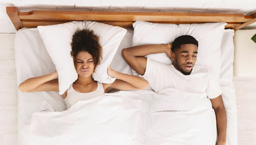 Couple in bed, man is snorin, woman is pressing pillow to her ears